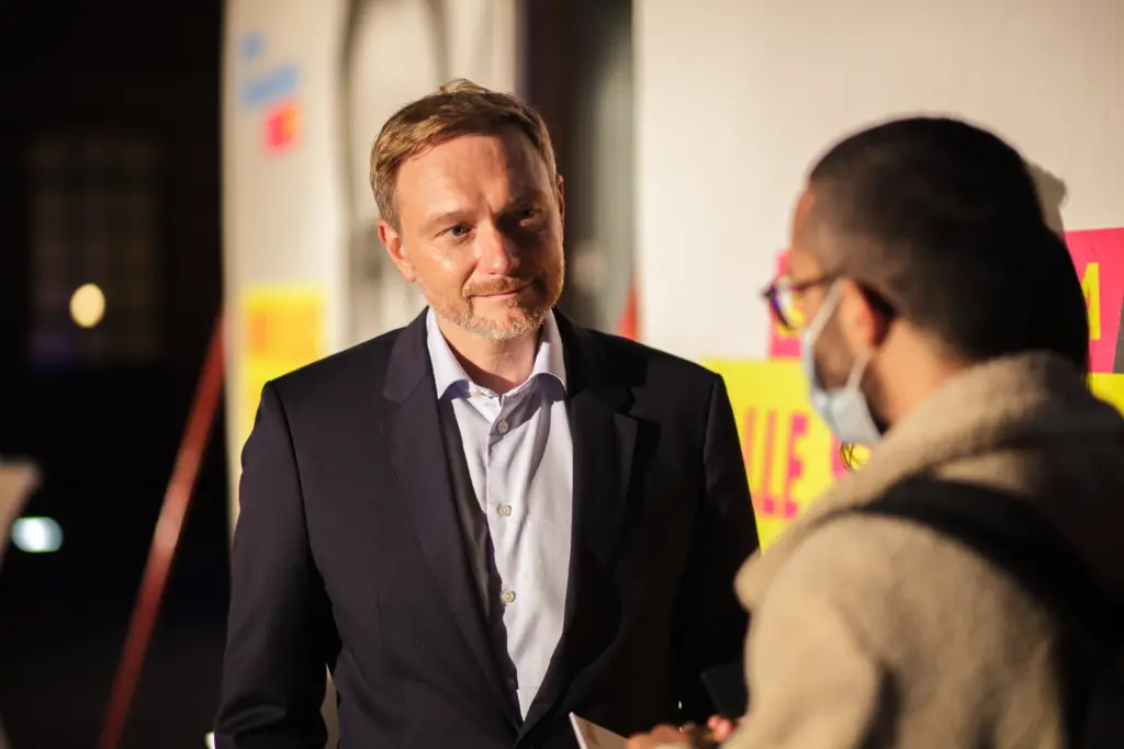 Christian Lindner steht in der Bildmitte vor Wahlplakaten, rechts stehen die beiden Redakteur*innen von Politikorange.