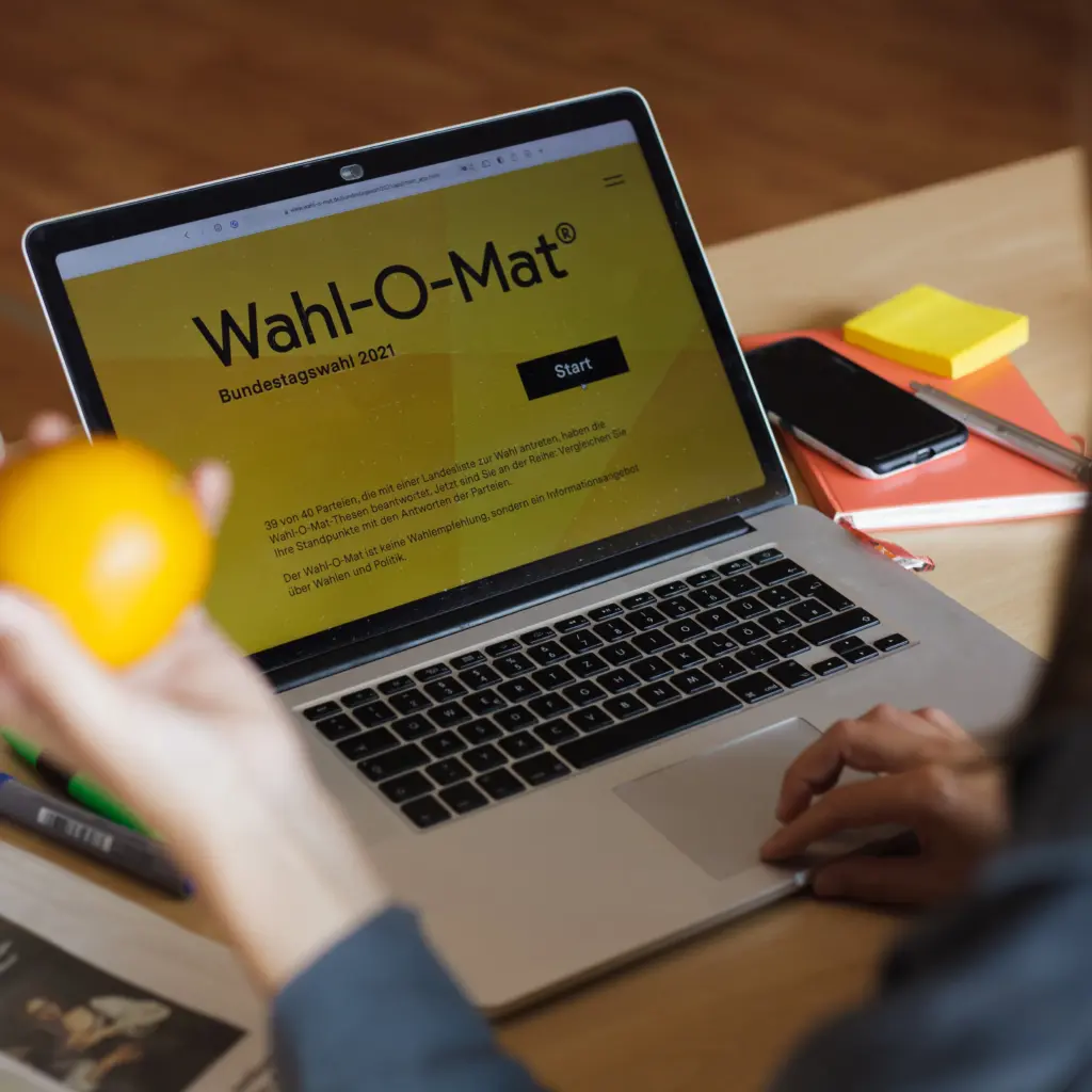 Ein Laptop auf einem Schreibtisch. Der Laptop zeigt den "Wahl-O-Mat". Im Vordergrund hält eine Hand eine Orange.