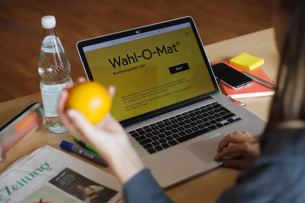Ein Laptop auf einem Schreibtisch. Der Laptop zeigt den "Wahl-O-Mat". Im Vordergrund hält eine Hand eine Orange.