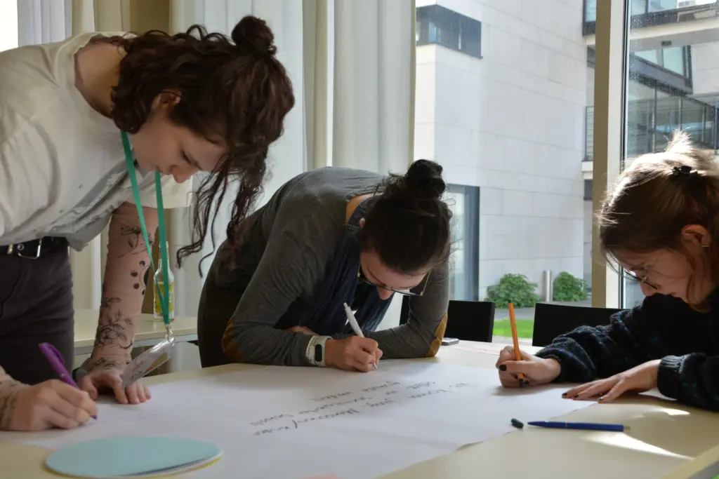 Drei junge Frauen schreiben auf einem Plakat ihre Forderungen nieder.